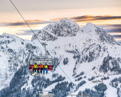 Skilift mit Fahrgästen, verschneite Berge und Sonnenaufgang im Hintergrund.
