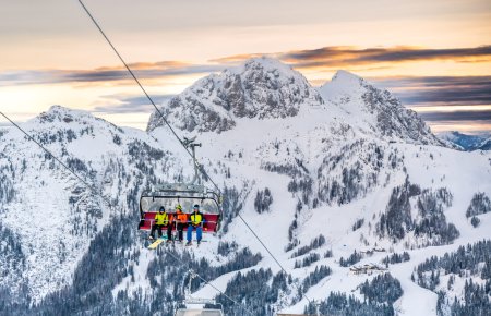 Skilift mit Fahrgästen, verschneite Berge und Sonnenaufgang im Hintergrund.