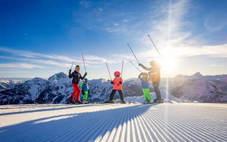 Skiers and children stand in the snow, sun behind the mountains.