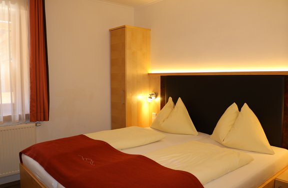 Hell beleuchtetes Schlafzimmer mit Doppelbett, roten Vorhängen und Holzschrank an der Wand