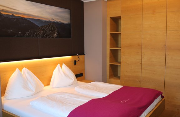 Doppelbett mit weißen Kissen und roter Decke vor Holzwand und Bergbild