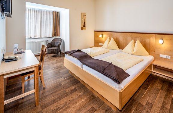 Hell eingerichtetes Hotelzimmer mit Doppelbett, Schreibtisch und Korbsessel am Fenster