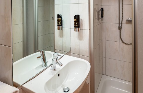 Kleines Badezimmer mit Waschbecken links und Duschkabine rechts, Spiegel und Seifenspender an der Wand
