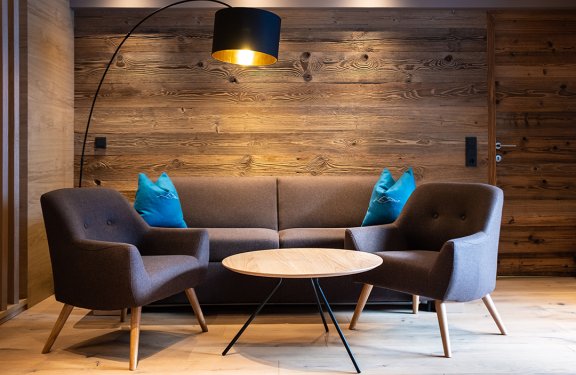 Gemütliche Sitzecke mit braunem Sofa, zwei Sesseln, blauem Kissen und runden Holztisch vor Holzwand