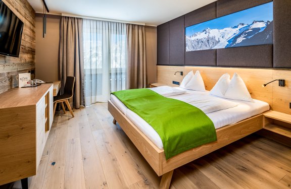 Modernes Hotelzimmer mit Doppelbett, grünem Überwurf, Holzmöbeln und Bergbild über dem Kopfteil