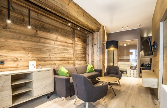 Gemütlicher Wohnbereich mit Holzwänden, Sofa, Sesseln, runden Tisch und Sauna im Hintergrund