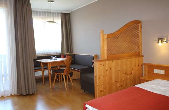 Helles Hotelzimmer mit Holzmöbeln, Sitzecke, Tisch und Bett mit roter Decke