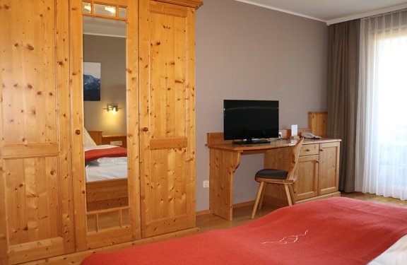 Schlafzimmer mit Holzkleiderschrank, Spiegel, Schreibtisch mit Fernseher und roter Bettdecke