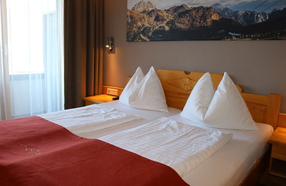Hell eingerichtetes Schlafzimmer mit Doppelbett, weißen Kissen und roter Decke vor Fenster und Bergbild