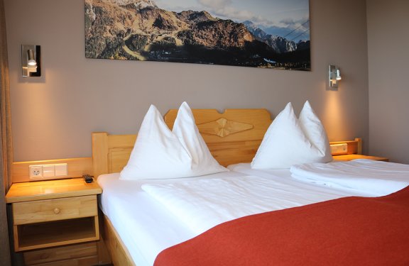 Doppelbett mit weißen Kissen und roter Decke, Holznachttische und Bergbild an der Wand