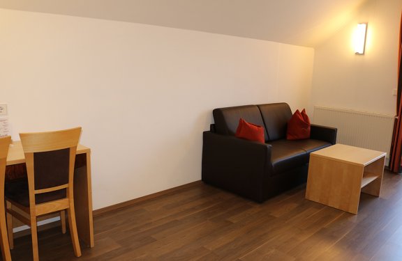 Kleines Zimmer mit braunem Sofa, roten Kissen, Holztisch und Stuhl vor weißer Wand