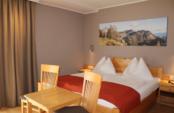 Hell eingerichtetes Schlafzimmer mit Holzbetten, roter Decke, Tisch und Stühlen vor braunen Vorhängen
