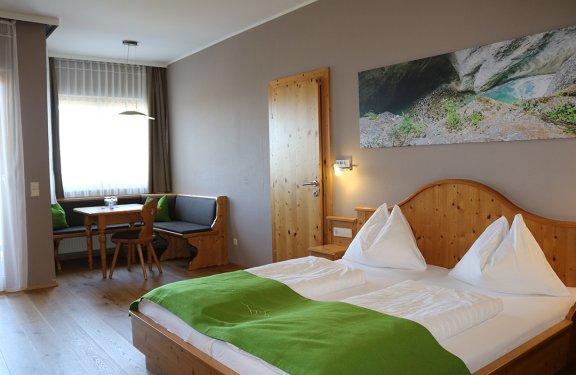 Helles Hotelzimmer mit Holzmöbeln, Doppelbett mit grünem Überwurf und Sitzecke am Fenster