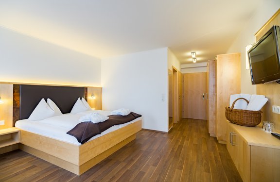 Hell eingerichtetes Hotelzimmer mit Doppelbett links und Fernseher rechts an der Wand