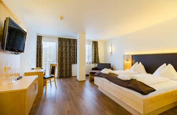 Helles Hotelzimmer mit Doppelbett, braunen Vorhängen, Fernseher an der Wand und Schreibtisch mit Stuhl