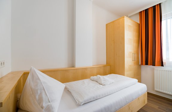 Einzelzimmer mit hellem Holzbett und orangefarbenem Vorhang